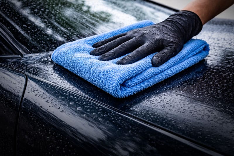 A hand using a microfiber towel to gently dry a wet car surface, demonstrating safe detailing technique to prevent swirl marks and preserve a clean, streak-free finish