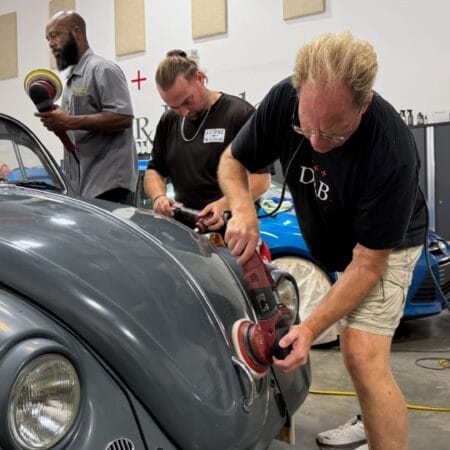 Mike Phillips demonstrates machine polishing on a gray Volkswagen Beetle while students practice beside him.