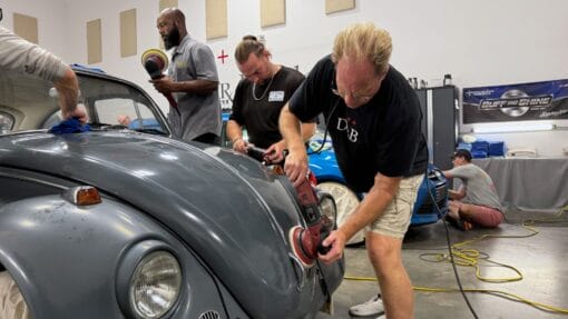Mike Phillips demonstrates machine polishing on a gray Volkswagen Beetle while students practice beside him.