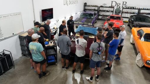 Mike Phillips leads a detailing lesson with students gathered around a burgundy Corvette in the training shop.