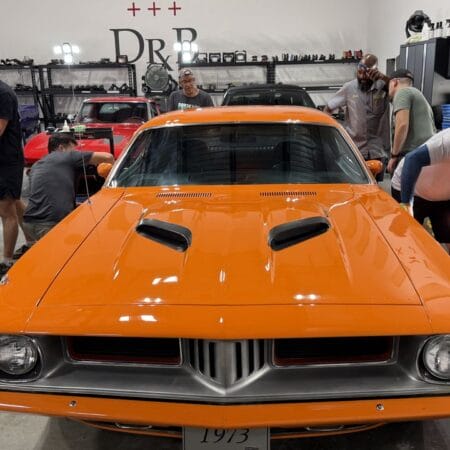 Students inspect the glossy paint on an orange Plymouth Cuda using a handheld inspection light.