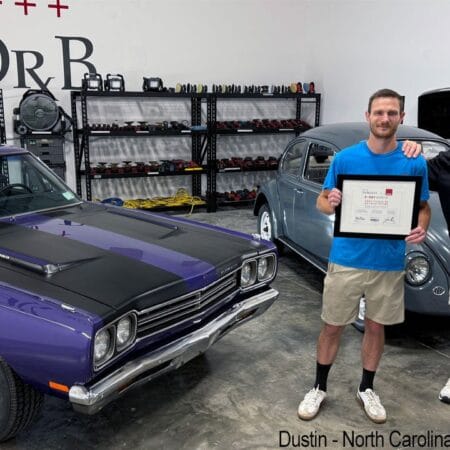 Mike Phillips stands with a student holding his detailing class certificate beside a purple Plymouth Road Runner.