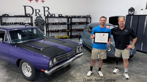 Mike Phillips stands with a student holding his detailing class certificate beside a purple Plymouth Road Runner.