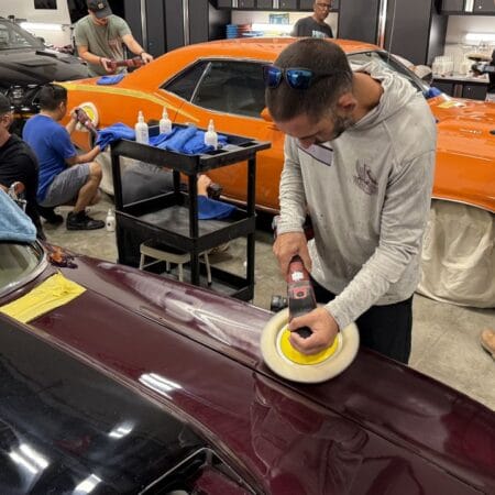 A student practices rotary polishing on a burgundy Corvette while classmates work on an orange Plymouth Cuda.
