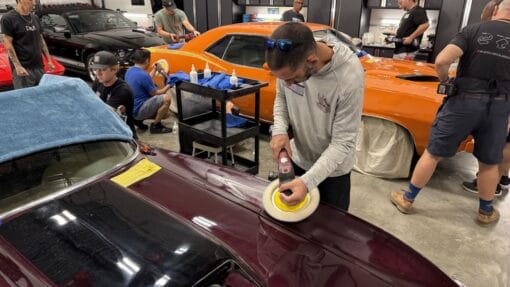 A student practices rotary polishing on a burgundy Corvette while classmates work on an orange Plymouth Cuda.