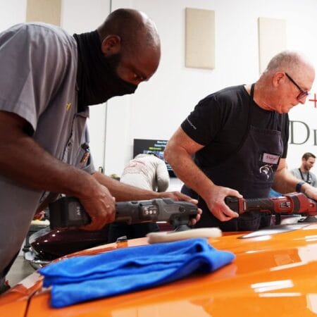 Two students practice with different machine polishers on the hood of an orange Plymouth Cuda.