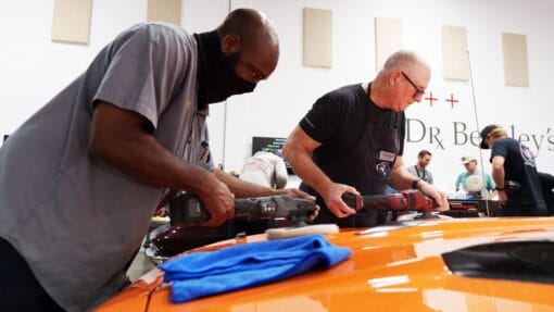 Two students practice with different machine polishers on the hood of an orange Plymouth Cuda.