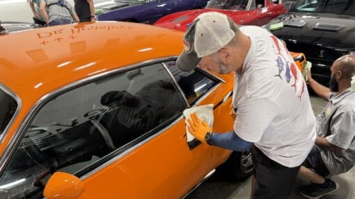 Students wearing gloves apply coating to the side panels of an orange Plymouth Cuda.