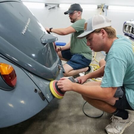 Students practice machine polishing the curved rear panels of a gray Volkswagen Beetle.