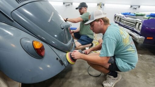 Students practice machine polishing the curved rear panels of a gray Volkswagen Beetle.