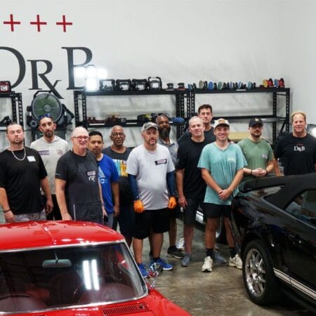 Mike Phillips and detailing students pose together behind a red Corvette and a black Ford Mustang in the training shop.