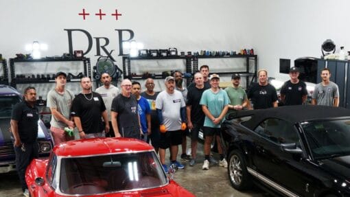 Mike Phillips and detailing students pose together behind a red Corvette and a black Ford Mustang in the training shop.